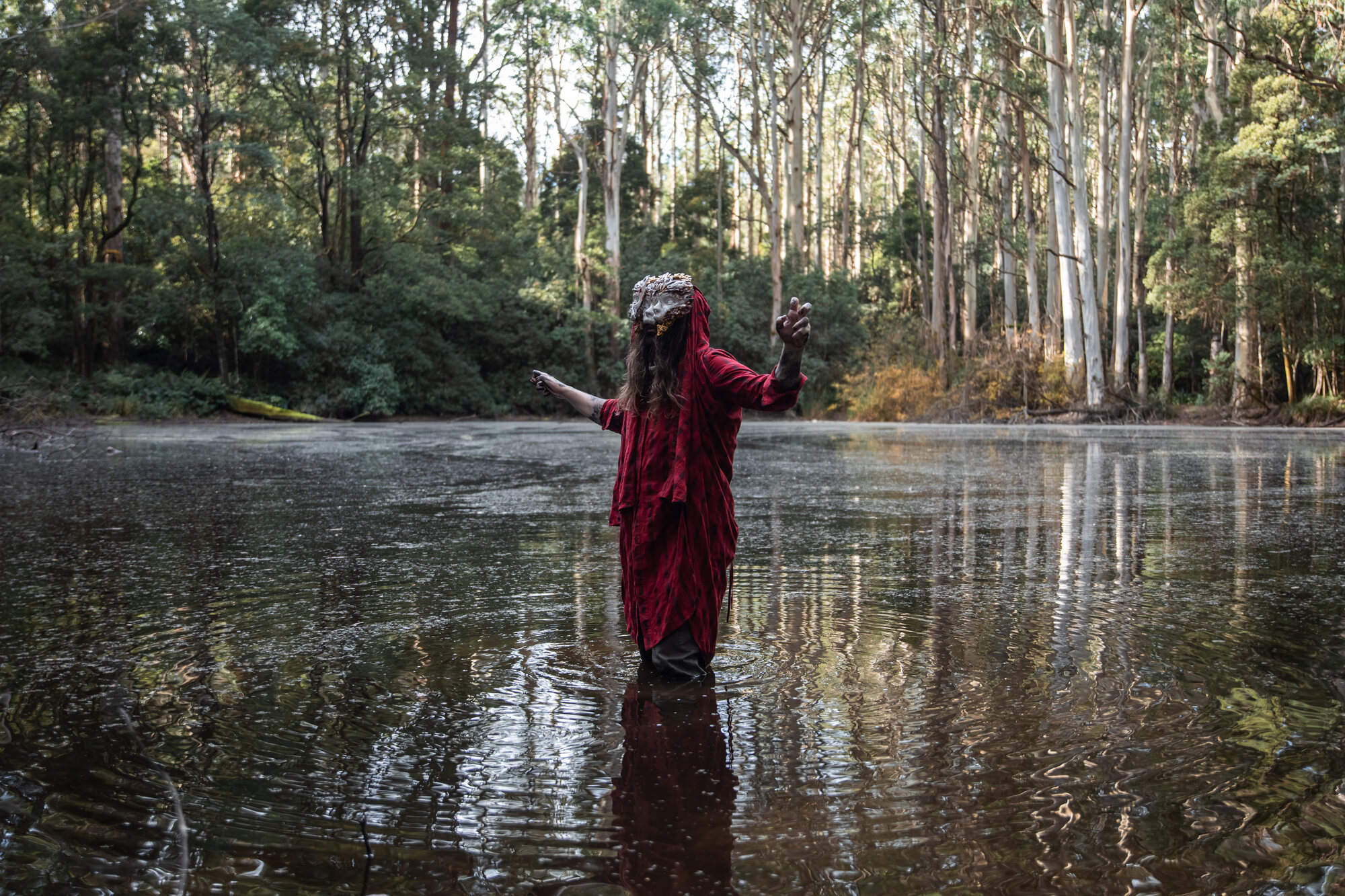 Wearable mask, red robed figure standing in still river, forest reflection